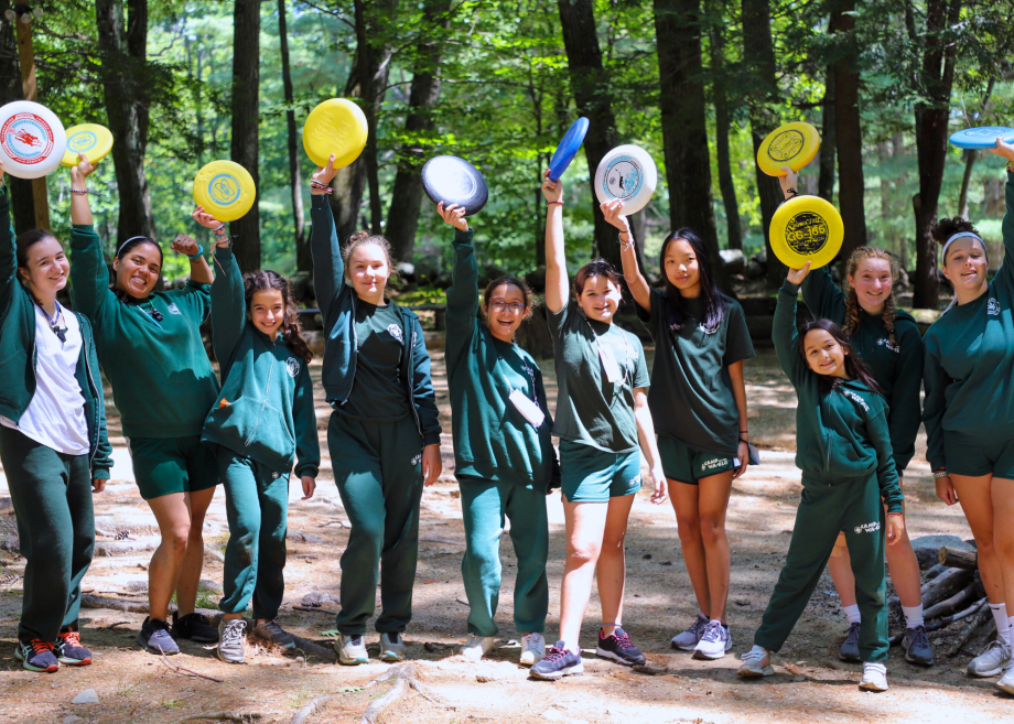 A large group of campers stand in a line, holding frisbees up in the air.